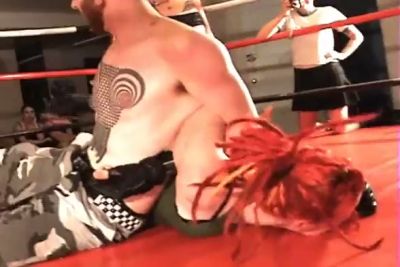 Black-haired dominatrix wields wooden paddle over kneeling opponent in red-walled ring - yesvids.com